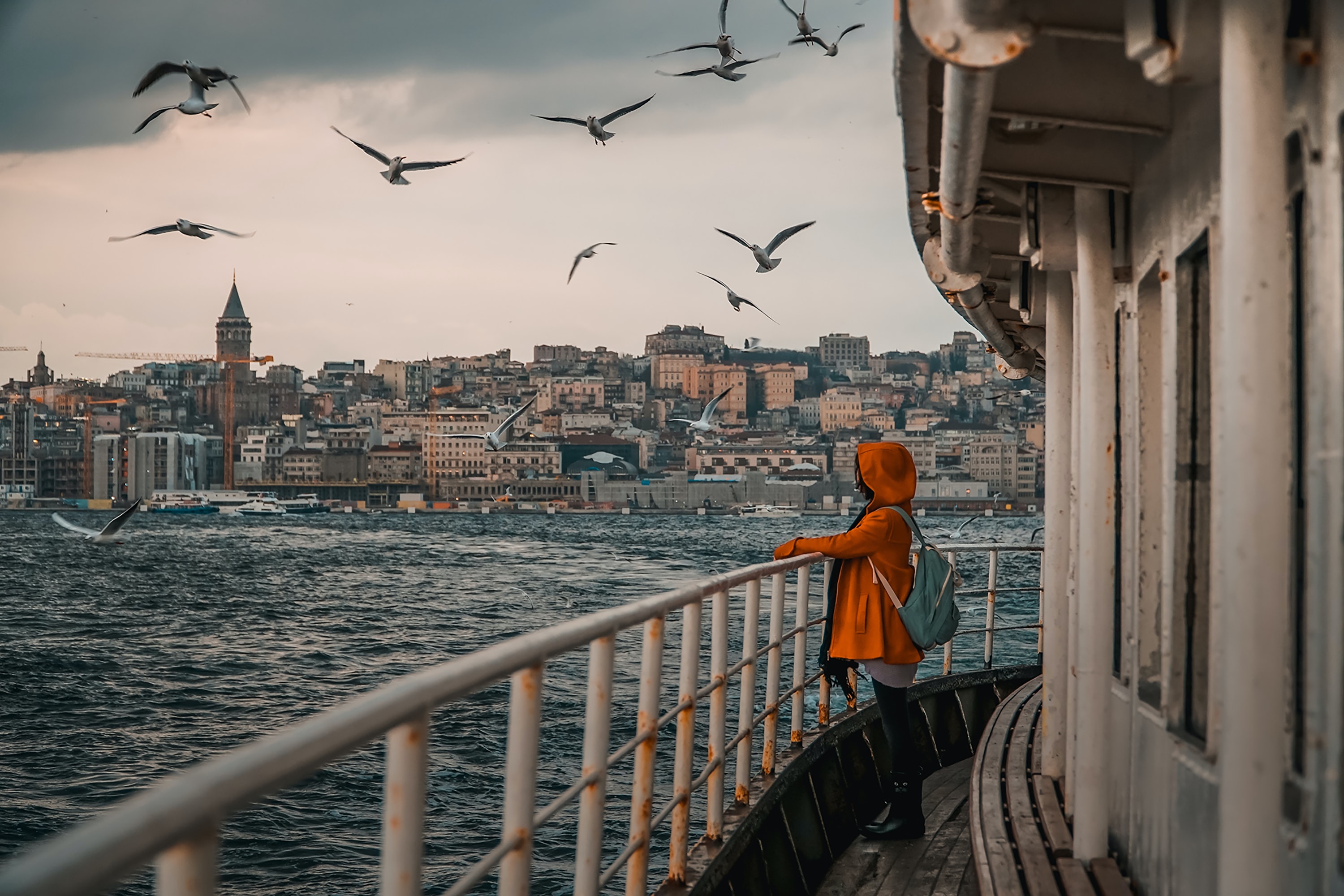 A woman wearing an orange rain coat and a backpack stands at the railing of a ferry crossing the Bosphorus on a cloudy day with Maiden's Tower in the background.