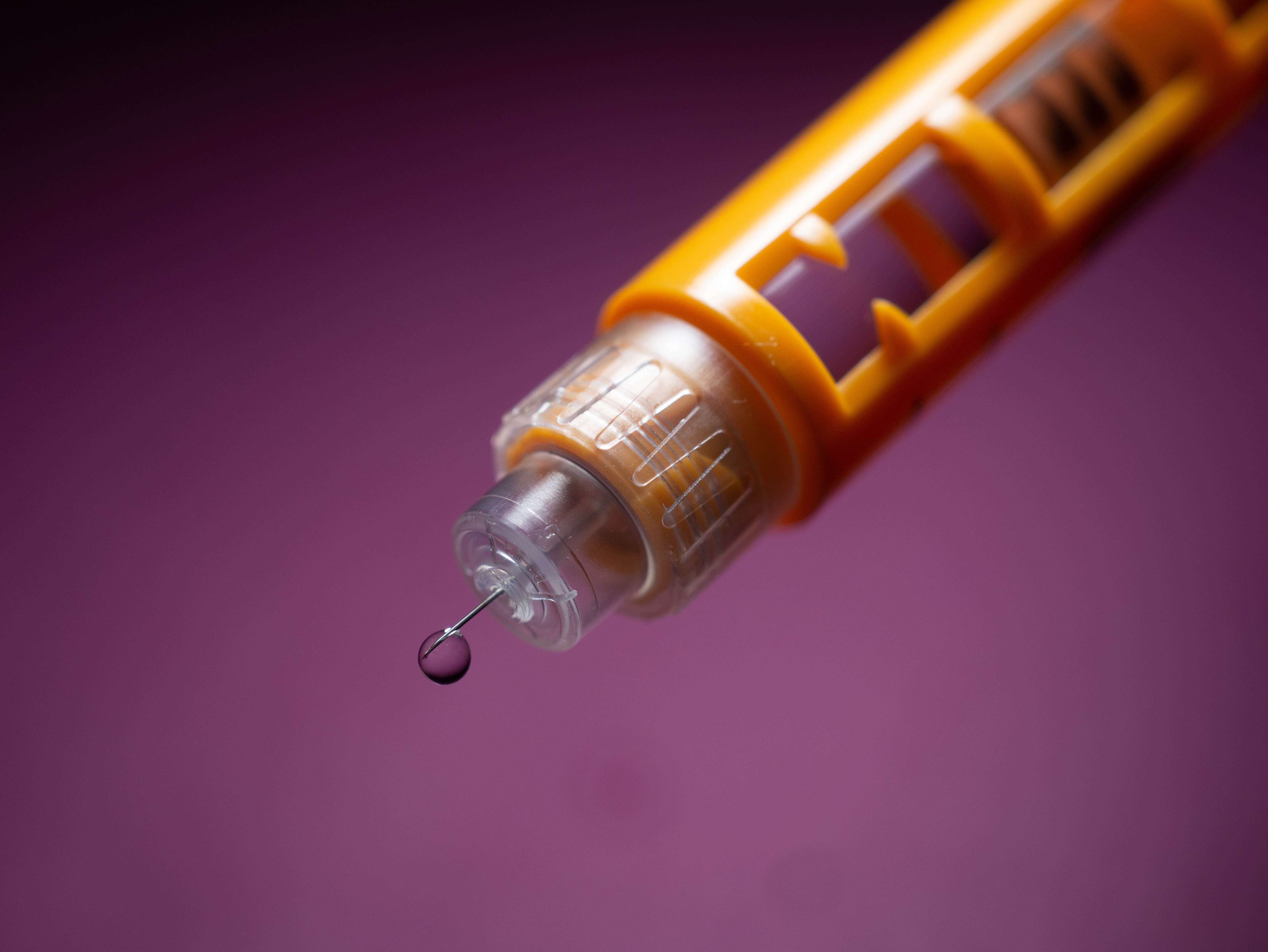 An orange insulin needle on a purple background with a drip of insulin coming out of it.