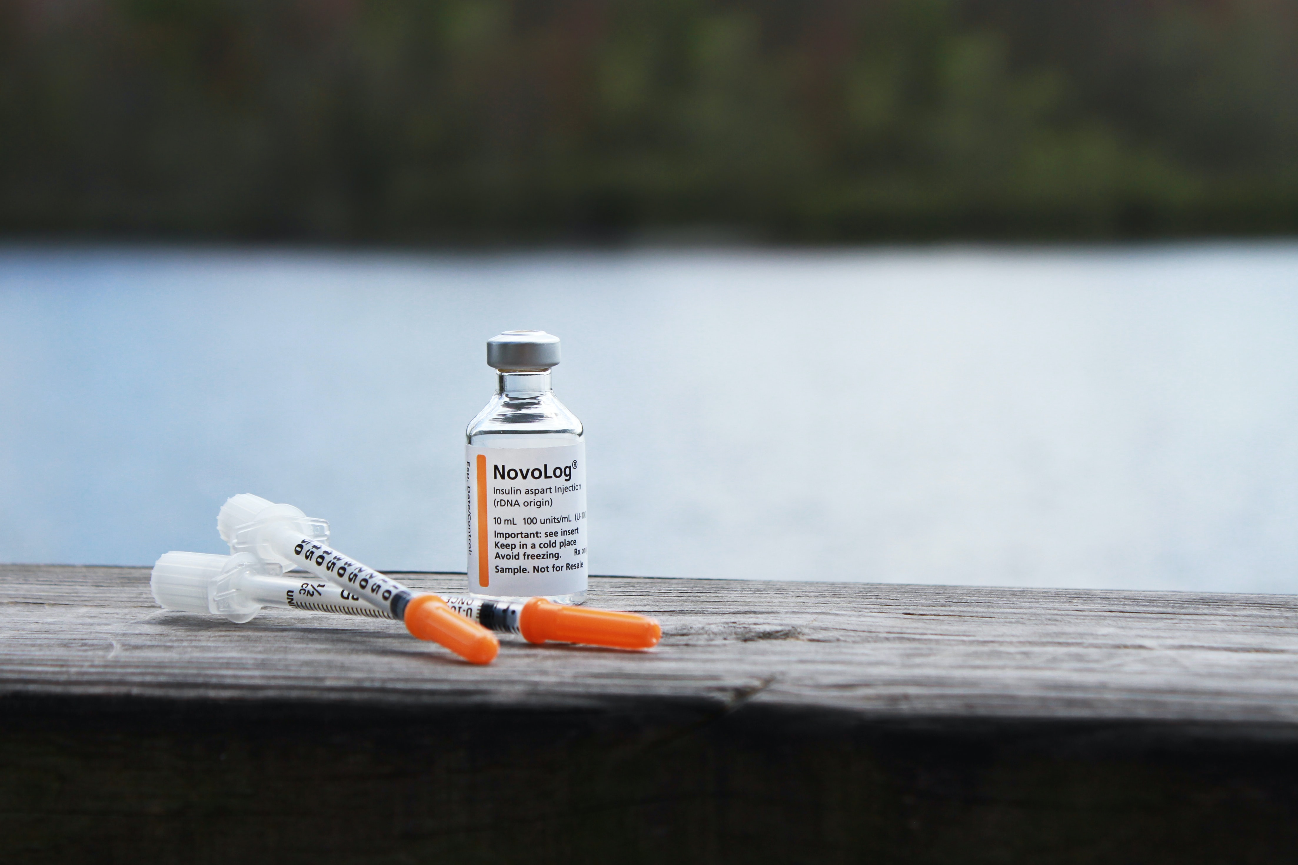 A vial of insulin sits on a wooden railing next to two needles with orange caps.