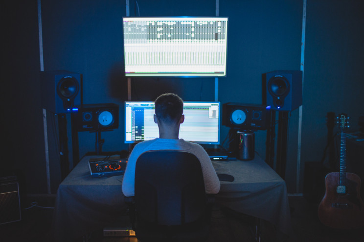 A person sits at a table with multiple speakers surrounding a giant screen with a music mixing program displayed on it.