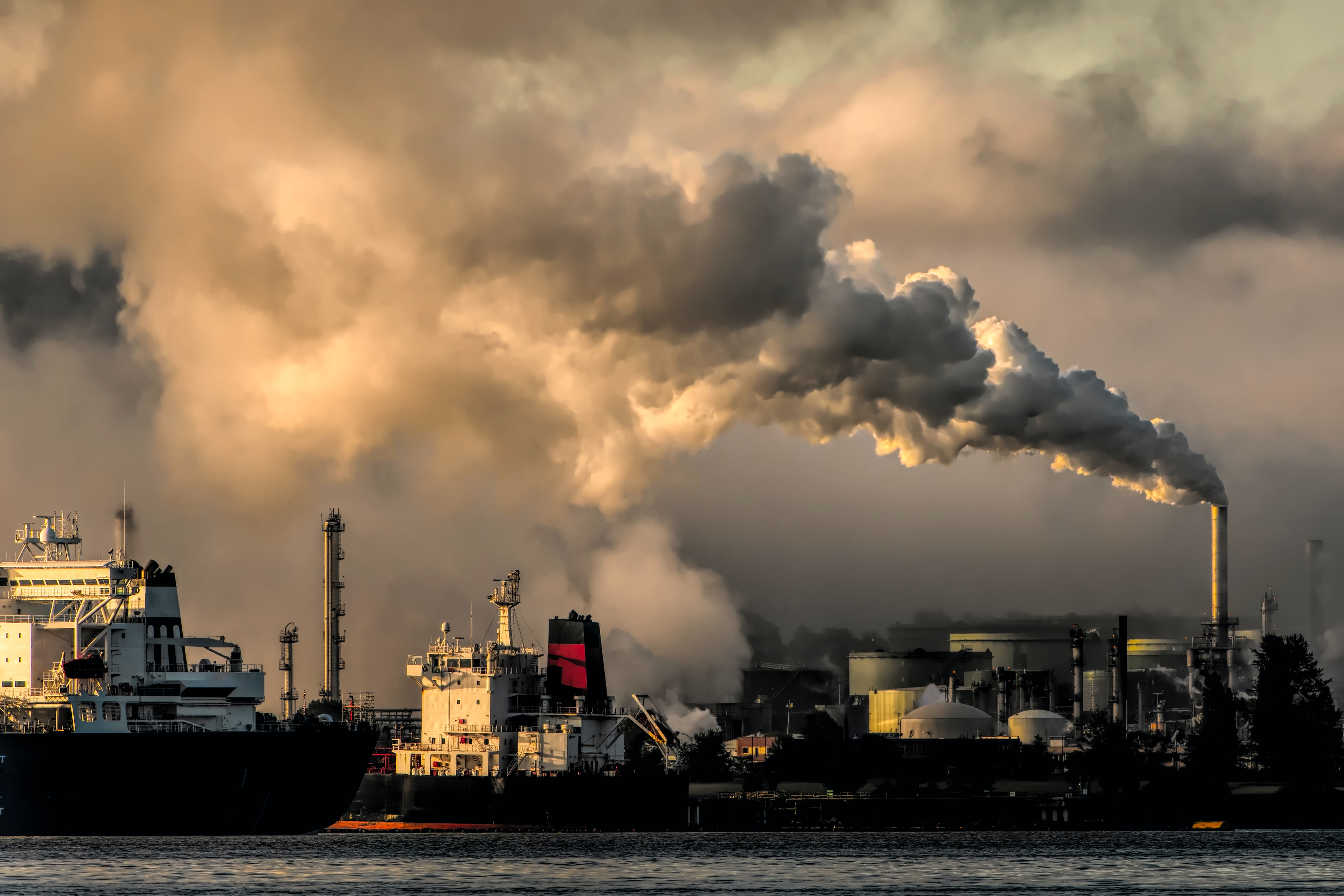 Factory towers emit billowing white and yellow clouds of smoke behind two large ships on the seaside.