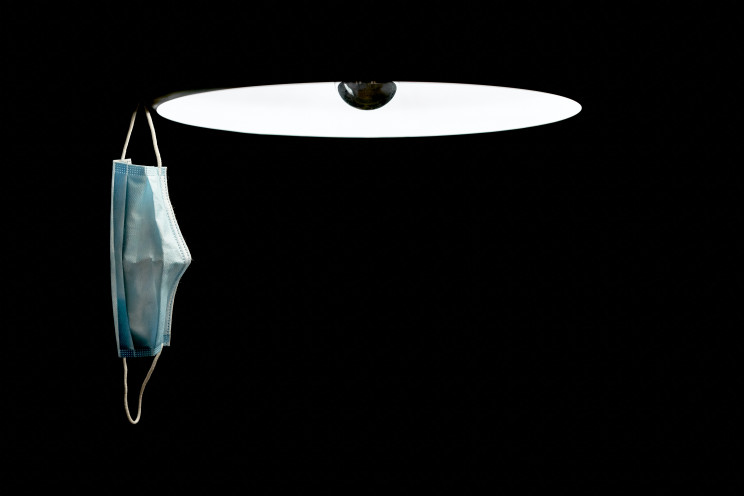 A COVID-19 mask hangs off of a lamp in a pitch black room.