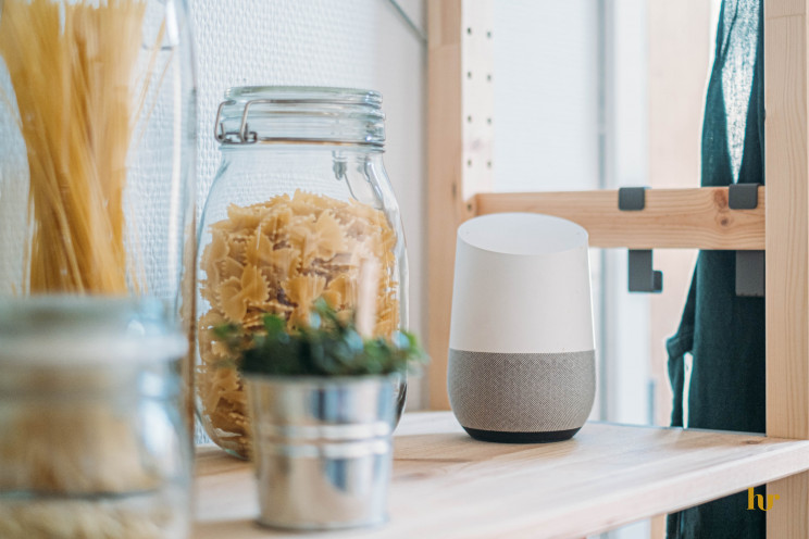 A Google Home smart speaker sits on a wooden shelf next to a jar filled with noodles in a kitchen.