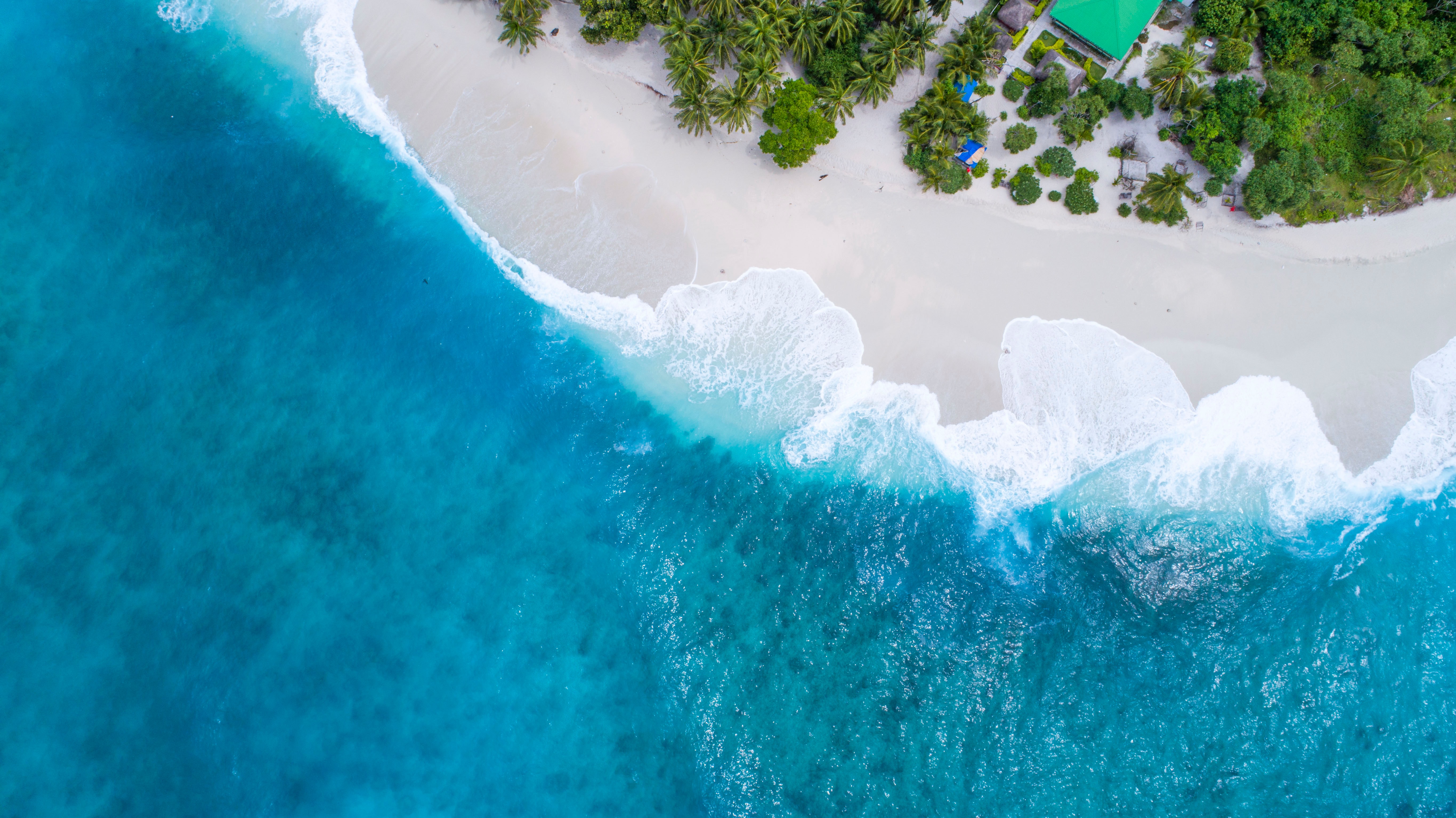 Deep blue waves come ashore near a house set amidst green trees in the Maldives.