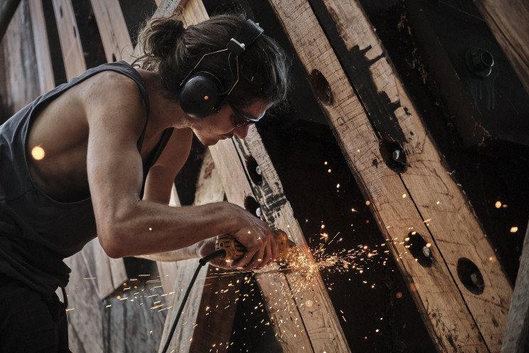 A worker with a ponytail using a tool to work on the wooden beams of the ship.